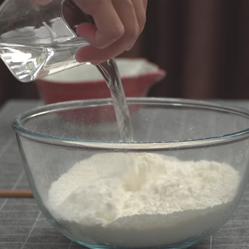 Bạn cho bột mì vào bát tô. Sau đó, bạn hòa tan 1 chút muối trong 130 ml nước ấm khoảng 60 °C.  Bạn bắt đầu đổ từ từ 100 ml nước vào bát bột, đồng thời dùng phới khuấy đều. Nếu thấy bột vẫn hơi khô thì cho tiếp dần dần từng thìa con nước vào để trộn đến khi không còn bột khô. Bạn chú ý không đổ quá nhiều nước cùng 1 lúc.  Bạn đổ bột ra mặt phẳng rộng để nhồi bột trong khoảng 15 phút. Cách nhồi cũng tương tự như khi nhồi bột bánh mì. Khi thấy khối bột càng ngày càng nhẹ tay hơn, lớp bột sờ vào nhẵn đều, không bị ướt hay khô quá là được.  Sau đó, bạn dùng màng bọc thực phẩm đậy lại, để bột nghỉ trong vòng 30 phút.